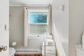 A bathroom with a toilet, a window with curtains, a towel rack with towels, and a heater. - Charming Lakefront Apartment with Deck, Patio and BBQ Grill (Orland)