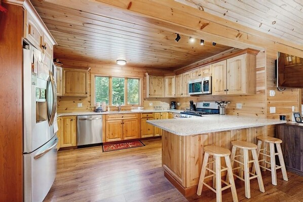 Ein Holzblockhaus-Interieur mit einer Küche, die Holzschränke, eine zentrale Kochinsel mit Barhocker, einen Edelstahlkühlschrank und eine Mikrowelle zeigt.