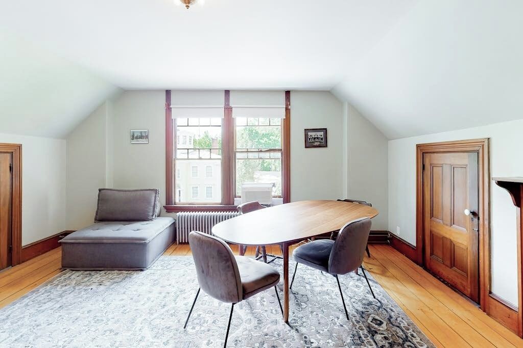 Een kamer met een houten tafel, twee stoelen, een grijze bank, een kamerbreed vloerkleed en een raam met uitzicht op gebouwen.