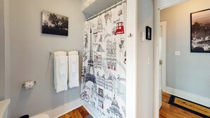Un baño con cortina de ducha adornada con ilustraciones de un paisaje urbano, un perchero con dos toallas, un cuadro enmarcado en la pared y un felpudo en la entrada.