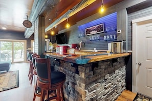 Una zona de bar con mostrador de piedra, taburetes, televisión y una estantería para vinos montada en la pared.