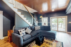 Una sala de estar con un sofá seccional gris, un perro negro, una escalera de madera y un ventanal grande.