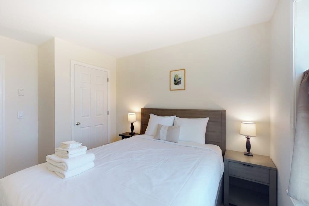 A hotel room with a neatly made bed, two bedside lamps, a door, and a small picture on the wall.