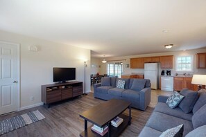 A living room with a sofa, a coffee table, and a TV. There is a kitchen in the background with wooden cabinets and a refrigerator.