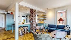 Ein modernes Wohnzimmer mit einem blauen Sofa, einem Korbstuhl, einem runden Couchtisch und einer Küchenzeile mit einer Ziegelwand.