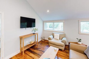 Ein Wohnzimmer mit Holzboden, einer Couch, einem Couchtisch, einem an der Wand befestigten Fernseher und einem Fenster.