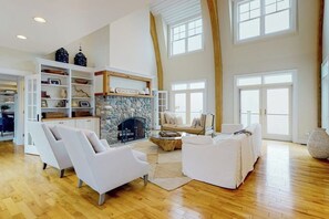A spacious living room with a stone fireplace, white sofas, and a high ceiling with large windows. - Spectacular oceanfront retreat with amazing views, sunroom & washer/dryer (Georgetown)