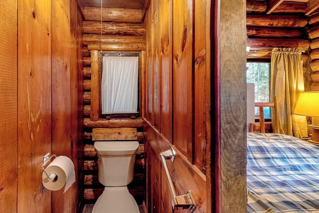 Houten blokhutinterieur met toilet, raam met gordijnen en bed met ruitbedekking.
