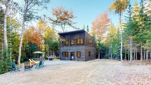 A modern log cabin with a dirt driveway, surrounded by trees and a fire pit area. - Contemporary retreat with grill, firepit, & patio - walk to beach (Lamoine)