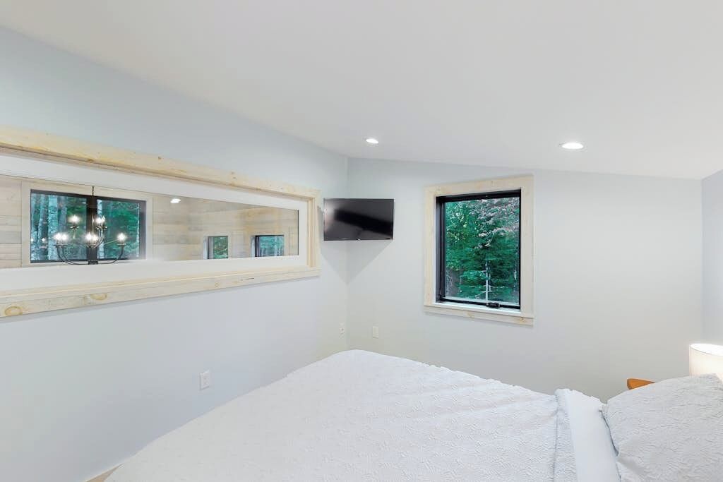 Un dormitorio con cama, un espejo grande, una ventana con vistas a árboles y luces empotradas en el techo.