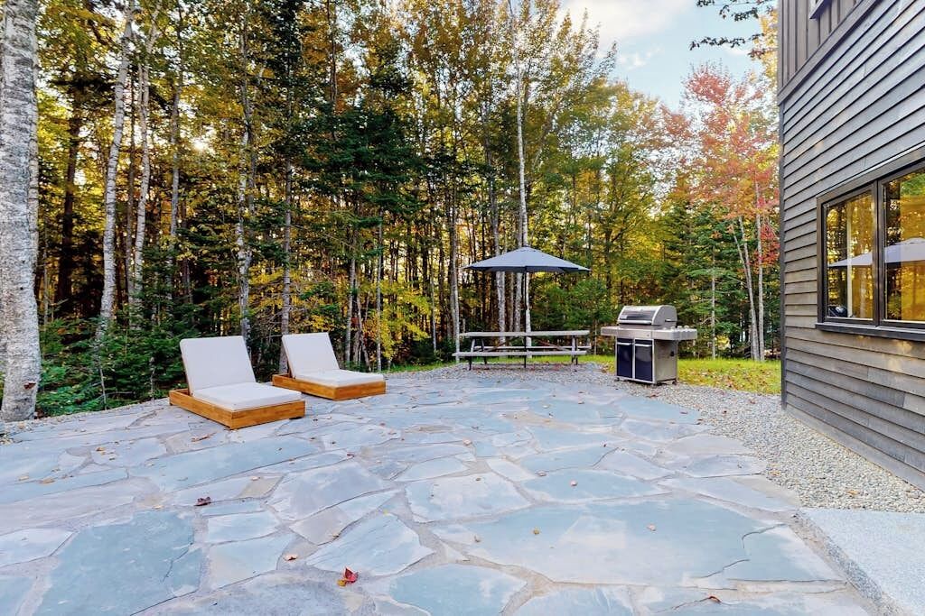 A stone patio with lounge chairs, a grill, and a gazebo in a forested area.