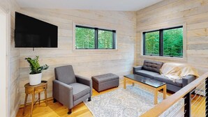 A modern living room with a sofa, armchair, ottoman, and coffee table, wooden walls, large windows, and a flat-screen TV. - Contemporary retreat with grill, firepit, & patio - walk to beach (Lamoine)