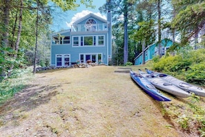 A two-story house with a front porch, a covered area with kayaks, and a stone wall. - Secluded waterfront home with firepit, gas grill, kayaks, dock, balcony, & patio (Phippsburg)