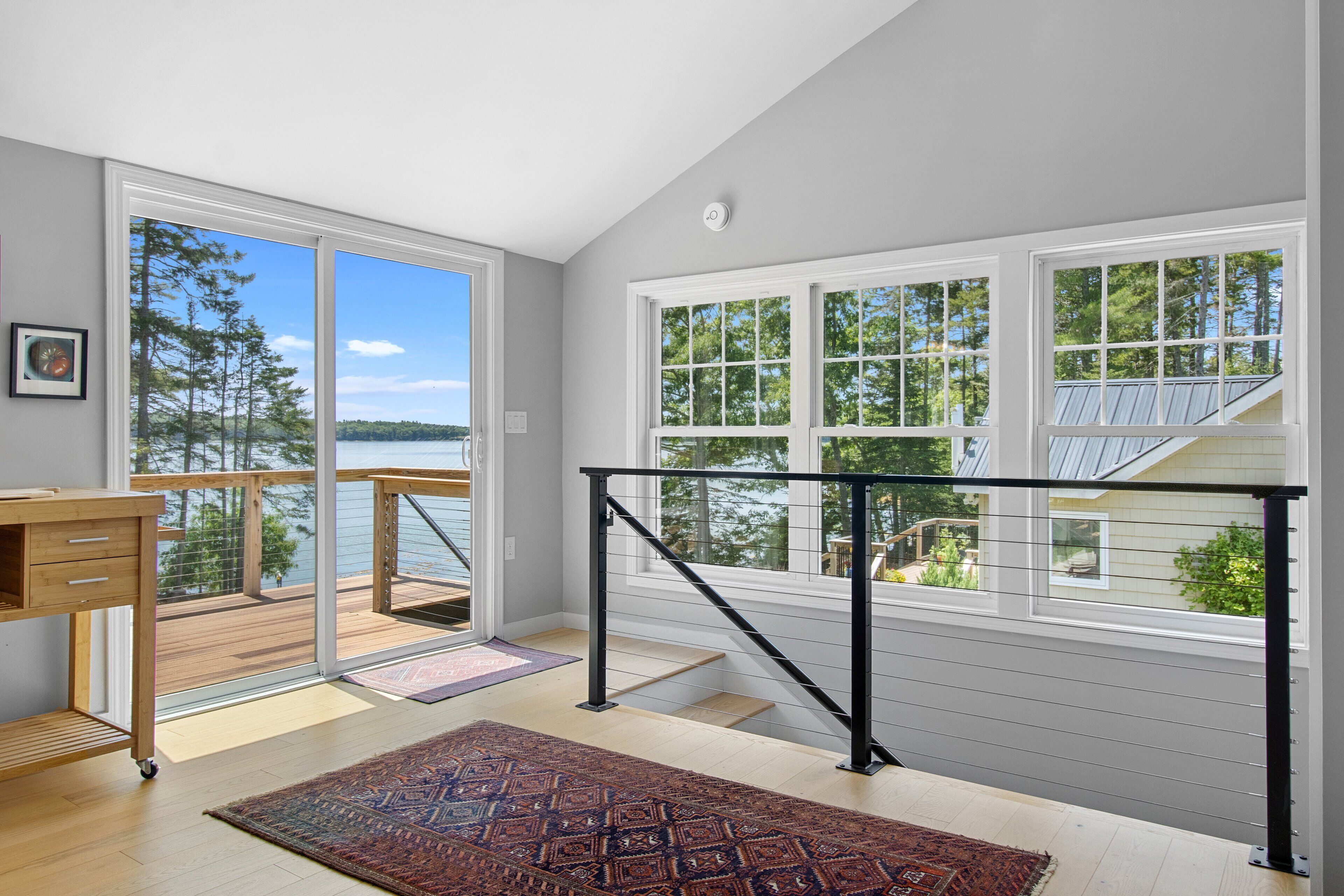 A room with a view of a lake, a wooden desk, a patterned rug, and a staircase with a railing.