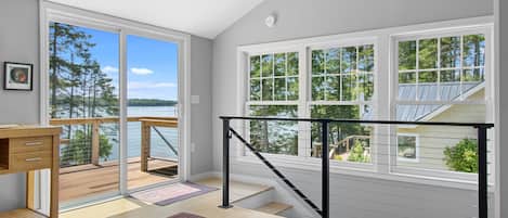 A room with a view of a lake, a wooden desk, a patterned rug, and a staircase with a railing.