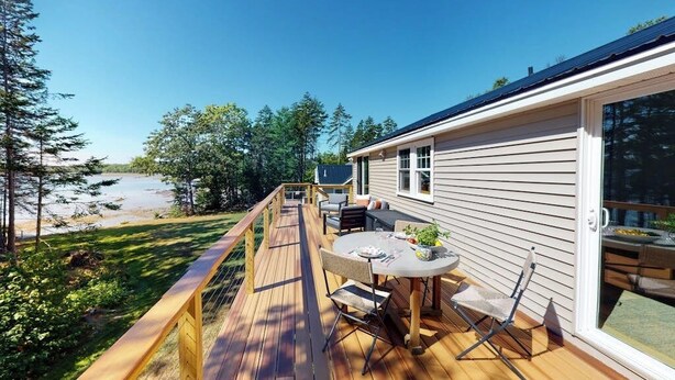 Um terraço com mobiliário de exterior, uma mesa e cadeiras, com vista para uma praia e uma floresta.