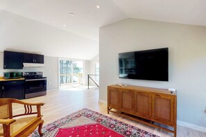 A modern living room with a flat-screen TV, wooden furniture, and a kitchen area with black cabinets and appliances.