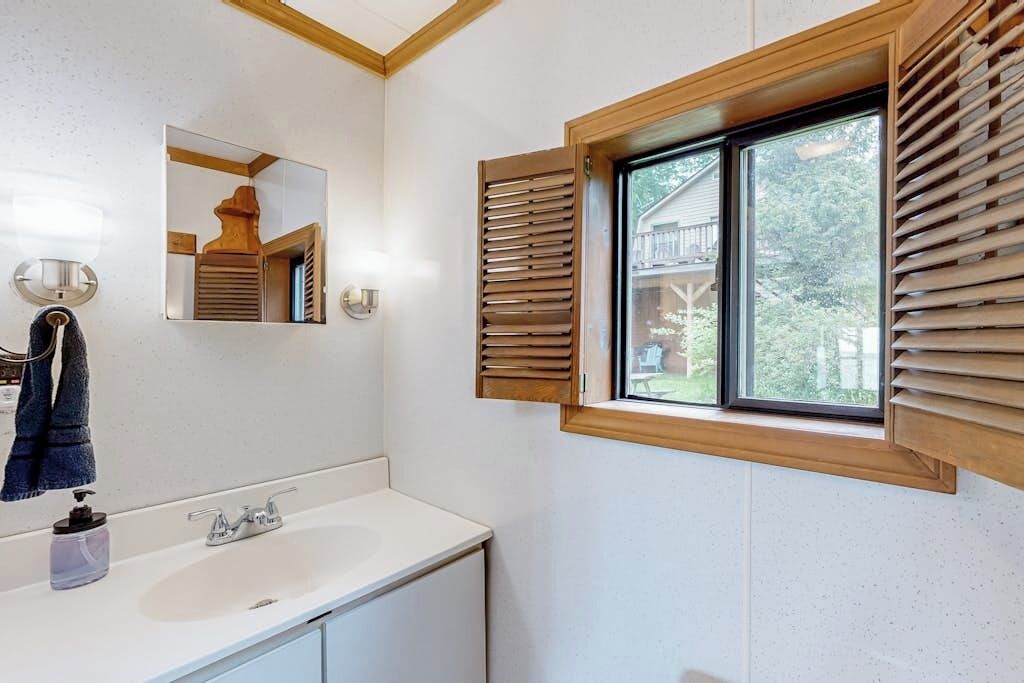 Une salle de bain avec un lavabo blanc, un miroir, des volets en bois sur une fenêtre et une serviette suspendue à un porte-serviettes.