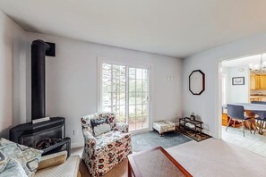 A living room with a fireplace, a floral armchair, a small ottoman, and a dining area with a table and chairs. - Idyllic waterfront home with furnished deck, grill, sweeping views & private W/D (Waldoboro)