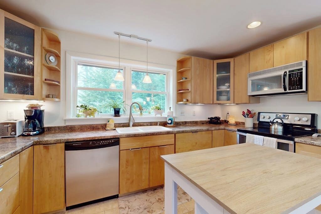 A modern kitchen with wooden cabinets, a dishwasher, a microwave, and a window with plants.
