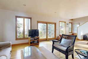 A spacious living room with a television, a sofa, a wicker chair, and large windows.