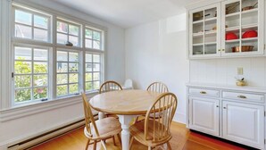 Un comedor con una mesa redonda y cuatro sillas, una ventana con múltiples divisiones y muebles de cocina blancos con puertas de vidrio.
