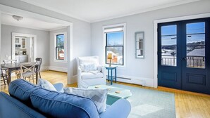 A modern living room with a sofa, a dining table, and a chair. There are two windows and a door. - Waterfront Duplex, walkable, harbor views, AC, dog friendly (Camden)