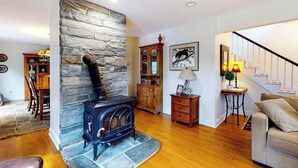 A living room with a stone wall, a wood-burning stove, a staircase, and a dining area. - Gorgeous water-view home & attached apartment (Camden)