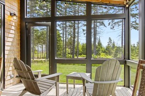 A wooden deck with two Adirondack chairs and a small table, overlooking a forested area through large windows. - Quiet, modern, dog-friendly home with ocean views, private trails & firepit (St. George)