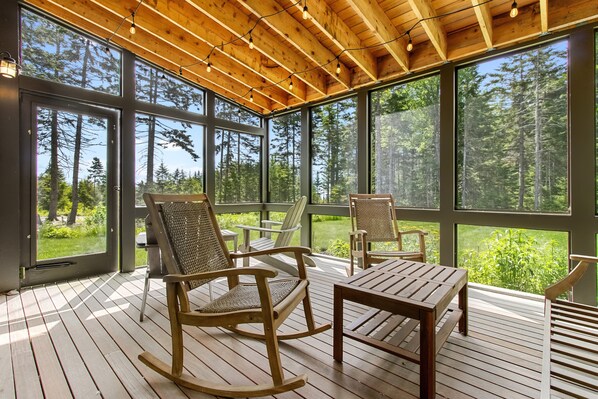 A wooden deck with rocking chairs and a table, large windows with forest views, and a wooden ceiling with exposed beams. - Quiet, modern, dog-friendly home with ocean views, private trails & firepit (St. George)
