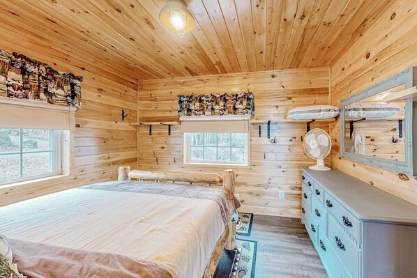 A wooden cabin bedroom with a bed, dresser, windows, and a fan. - Lake-view two-story home with beach access, wood stove, grill, W/D, & fast WiFi (Greenville)