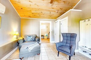 Een kamer met een grijze bank, een blauwe fauteuil, een houten plafond en een wit kastje met een plek voor dierenvoer.