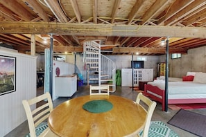Een kamer met een houten plafond, een spiraaltrap, een bed, een televisie en een eettafel met stoelen.