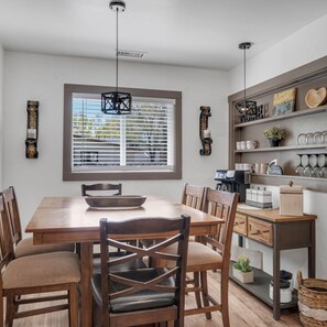 A dining area with a wooden table and chairs, a window with blinds, and a coffee station with mugs and a coffee maker. - WALK TO EVERYTHING, REMODELED, BEAUTIFUL NEIGHBORHOOD, LARGE YARD, AND FENCED! (Williams)