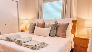 Cama bem arrumada com colcha estampada, travesseiros e uma toalha enrolada. Duas mesas de cabeceira com abajures, uma janela com persianas e piso de madeira.