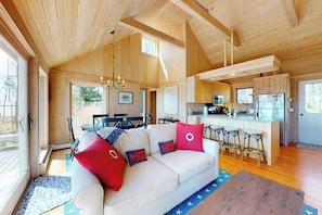 A cabin interior with a sofa, kitchen, dining area, and large windows. - Oceanfront retreat with sunrise views, screened porch, & deck (Brooklin)