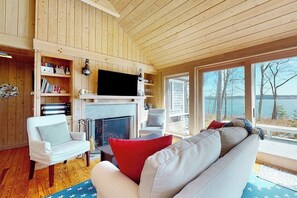 A cozy cabin interior with a fireplace, bookshelves, a television, and a view of the outdoors. - Oceanfront retreat with sunrise views, screened porch, & deck (Brooklin)