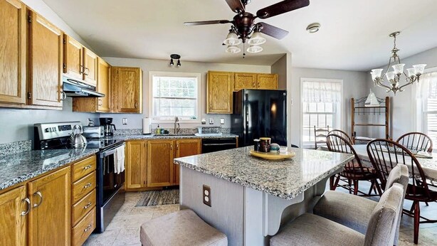 A kitchen with wooden cabinets, a black refrigerator, granite countertops, a ceiling fan, and a dining area with chairs. - Charming cottage near the beach with balcony, deck, grill, fireplace - dogs OK (Georgetown)