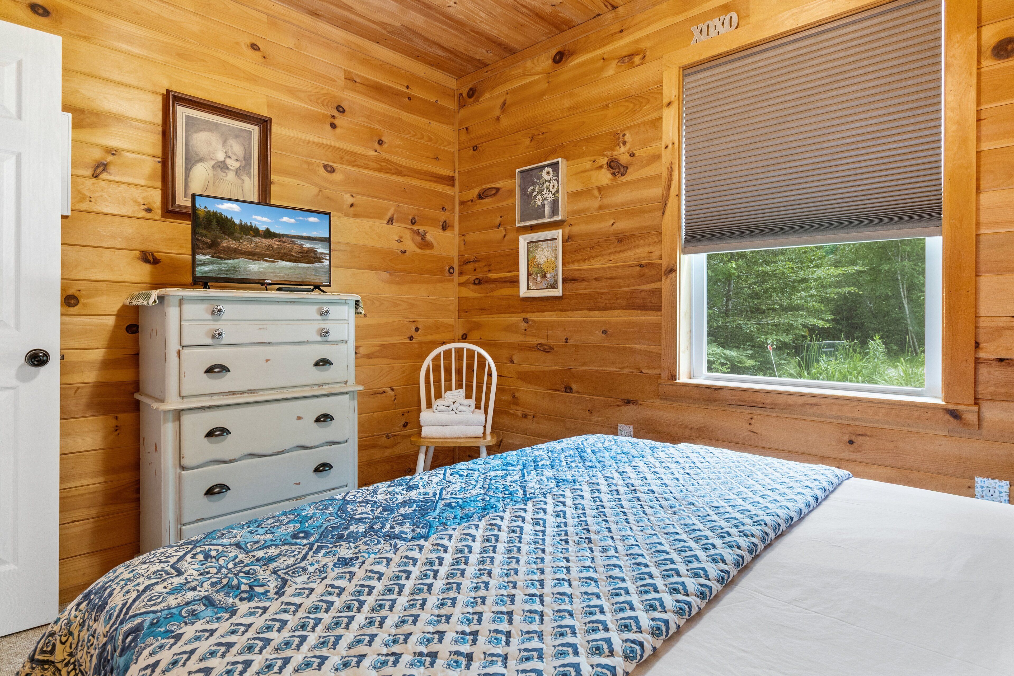 A wooden cabin room with a bed, dresser, chair, and a window with blinds.