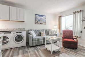 Un espace de vie compact comprenant un canapé à rayures, un fauteuil rouge, une table basse en verre, un lave-linge et un tableau représentant une scène de plage.