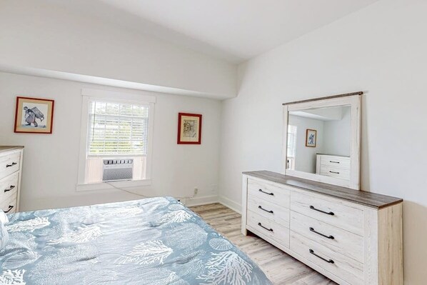 Ein Schlafzimmer mit Bett, Kommode, Spiegel, Fenster und zwei gerahmten Bildern an der Wand.