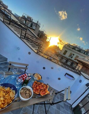 Una terraza en la azotea con una mesa lista para una comida, con vistas a la puesta de sol, una barandilla y un paisaje urbano de fondo.