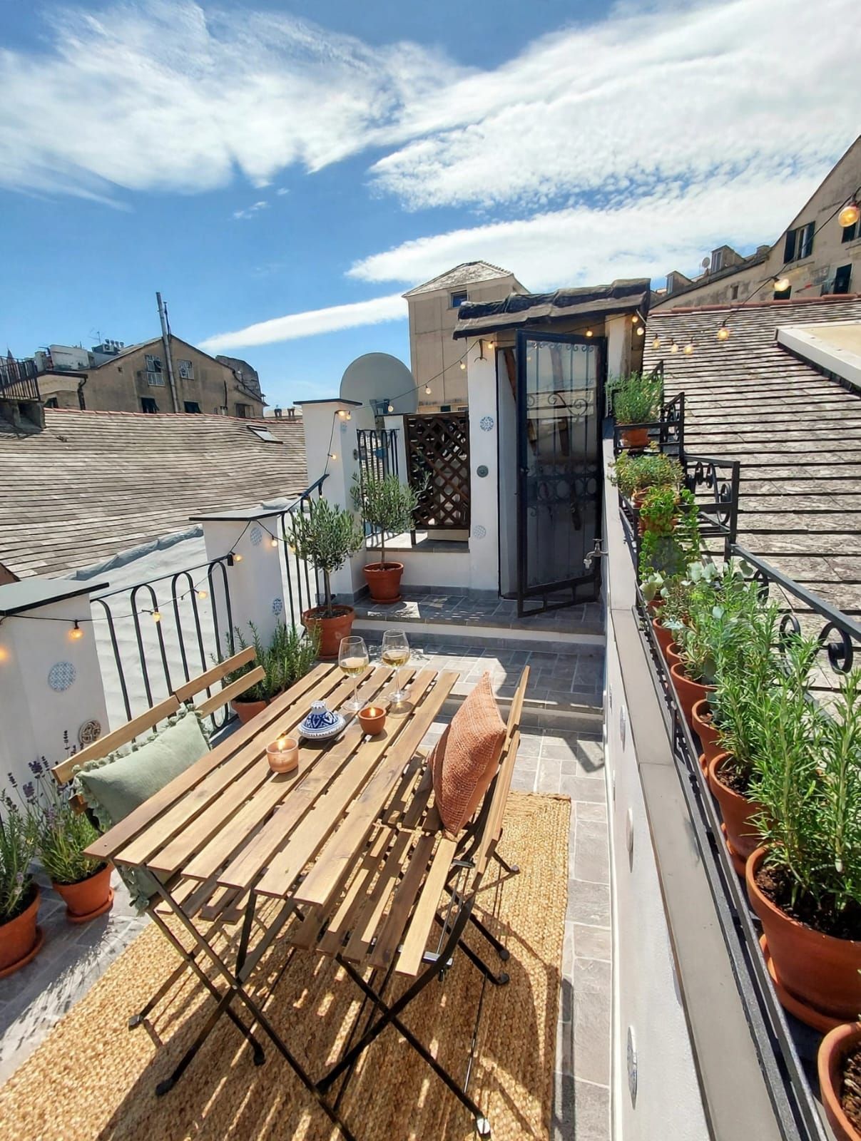 A rooftop terrace with a wooden table and chairs, potted plants, and a small seating area.