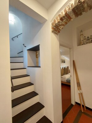 Eine Treppe mit schwarzen Stufen und weißen Wänden, eine Wand als Ziegelakzent und ein Bett mit einem gestreiften Kissen im angrenzenden Zimmer.
