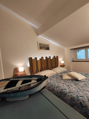Un dormitorio con cama, cabecera, mesita de noche, lámpara, cuadro y ventana.