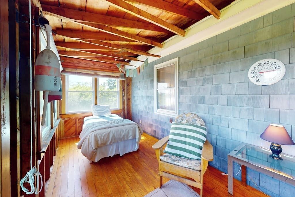 A room with a wooden ceiling, a bed, a chair, a table, and a wall clock.