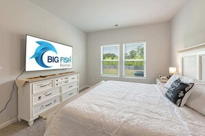 Un dormitorio con cama, cómoda, televisor mostrando 'BIG FISH Rentals' y ventana con persianas.
