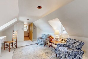 Ett rum i loftstil med ett sluttande tak, en trästol, en mönstrad fåtölj, ett litet bord med en lampa och utsikt genom en öppen dörr.