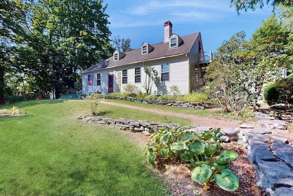 A two-story house with a stone pathway leading to the front door, surrounded by a well-maintained lawn and various shrubs. - Elegant riverside retreat with breathtaking views and modern comforts (Wiscasset)