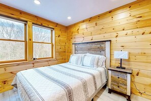 Een slaapkamer met houten lambrisering, een bed, nachtkastje, lamp en grote ramen.